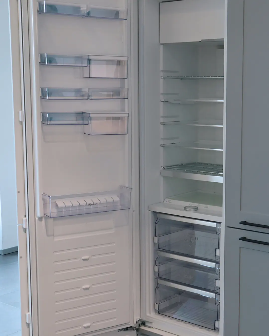 Geöffneter Einbaukühlschrank mit leeren Glasablagen und Schubladen in der grauen Häcker Landhausküche in G-Form