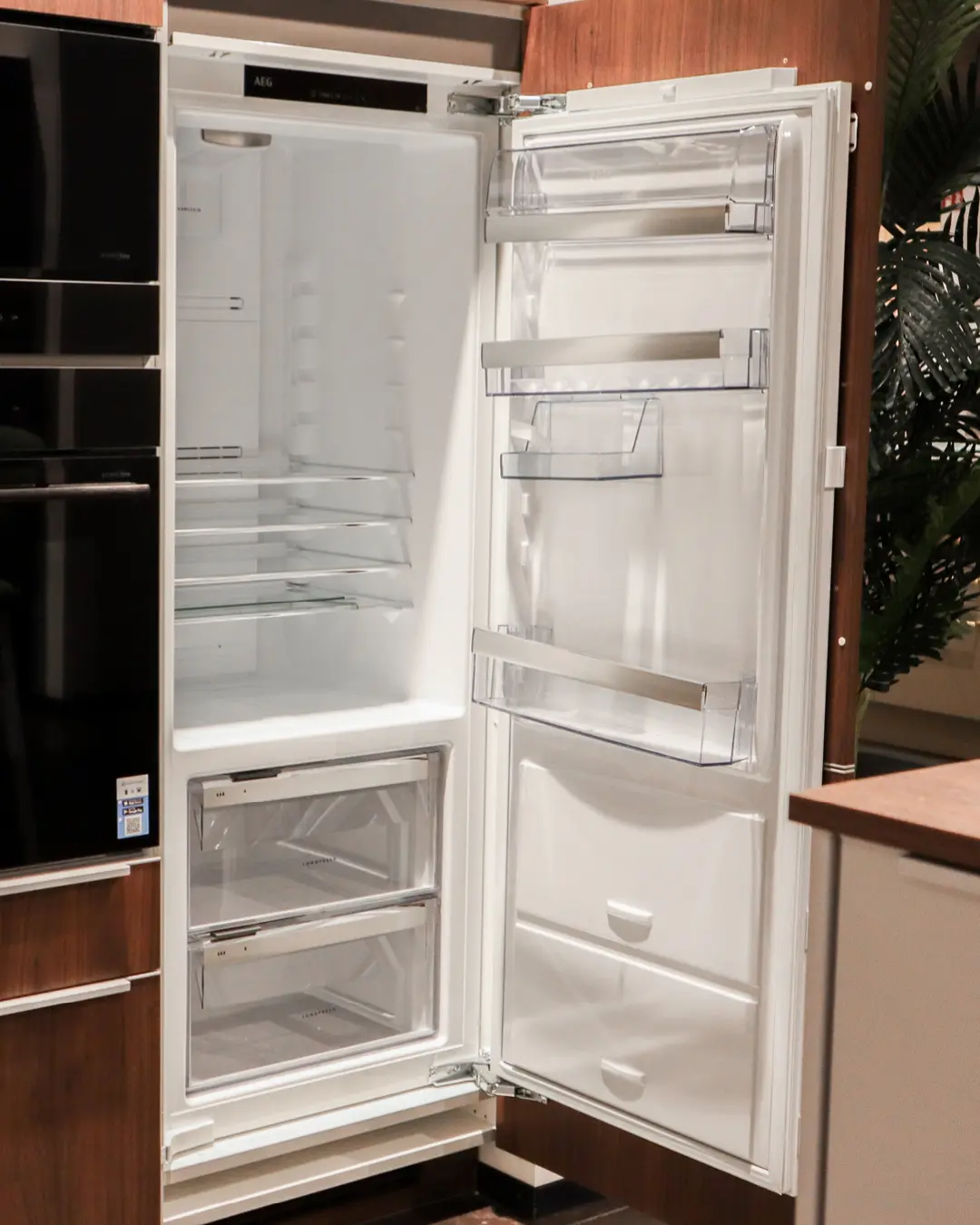 Geöffneter Einbaukühlschrank von AEG mit leeren Glasablagen in der Nobilia-Küche, daneben schwarze Einbaugeräte