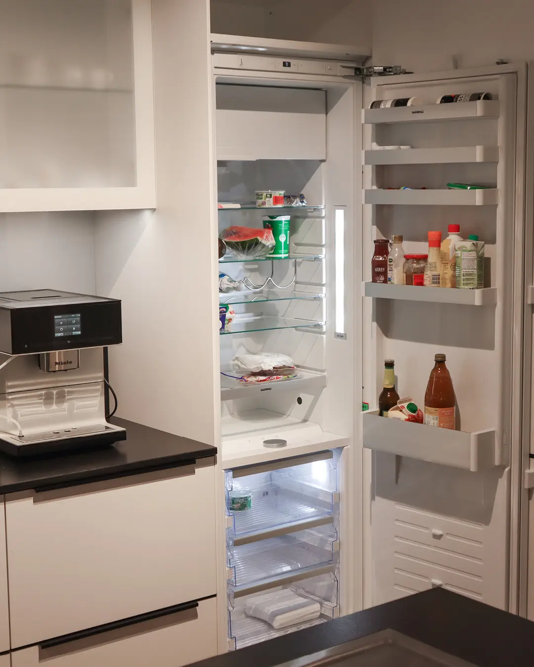 Geöffneter Einbaukühlschrank mit gefüllten Türfächern in kristallweißer Häcker Küche, daneben Kaffeevollautomat auf dunkler Arbeitsplatte