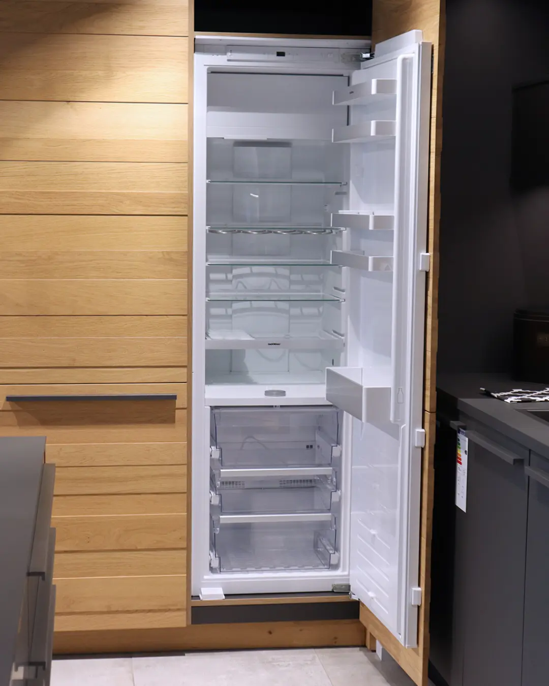 Geöffneter Einbaukühlschrank mit leeren Glasablagen in einer Leicht Küche mit Fronten in Holzoptik