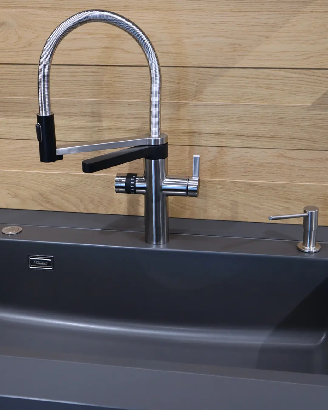 Detail der carbongrauen Leicht Küche: Edelstahl-Armatur mit Schlauchbrause an dunkler Spüle vor Wand in Holzoptik