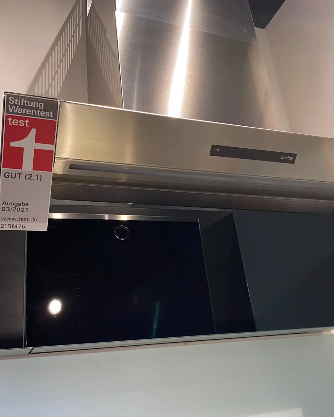 Edelstahl-Dunstabzugshaube von berbel über schwarzem Kochfeld in matt-weißer Nobilia Küche, mit Schild „Stiftung Warentest test GUT (2,1), Ausgabe 03/2021“