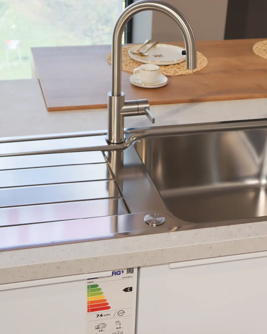 Detail der weiß-grauen Nobilia Küche in L-Form: Edelstahlspüle mit hohem Armatur und Holzablage mit Geschirr