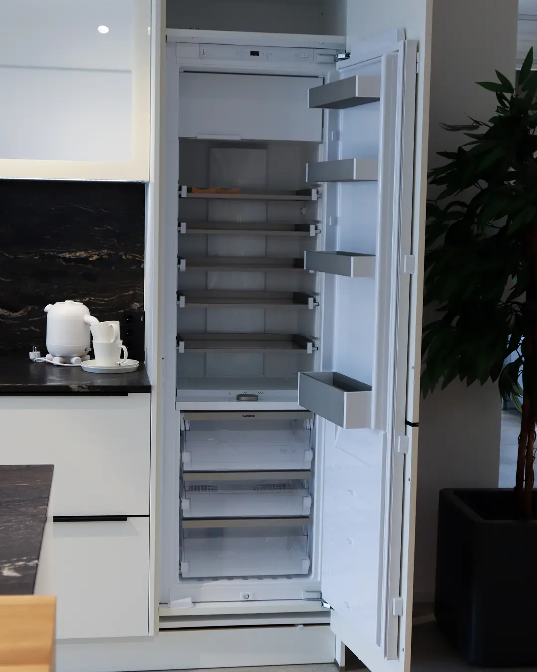 Geöffneter Einbaukühlschrank mit leeren Ablagen in der weißen Häcker Küche KBK17, daneben Arbeitsplatte in Steinoptik