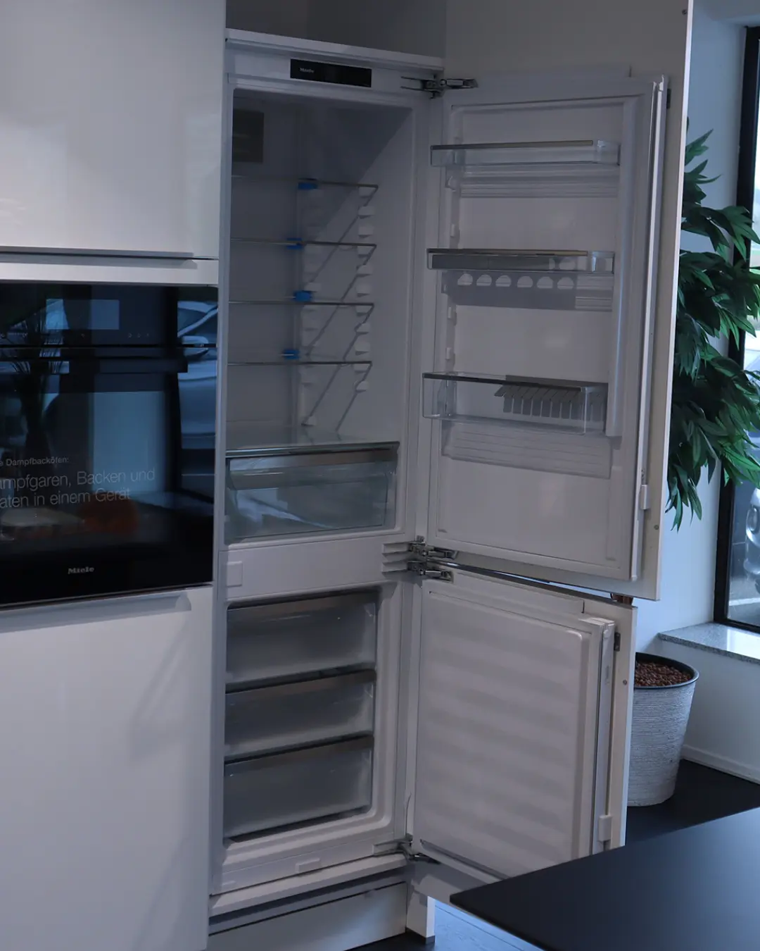 Geöffneter Einbaukühlschrank mit leeren Fächern in der weißen Hochglanz Küche von Häcker, daneben ein Einbaubackofen