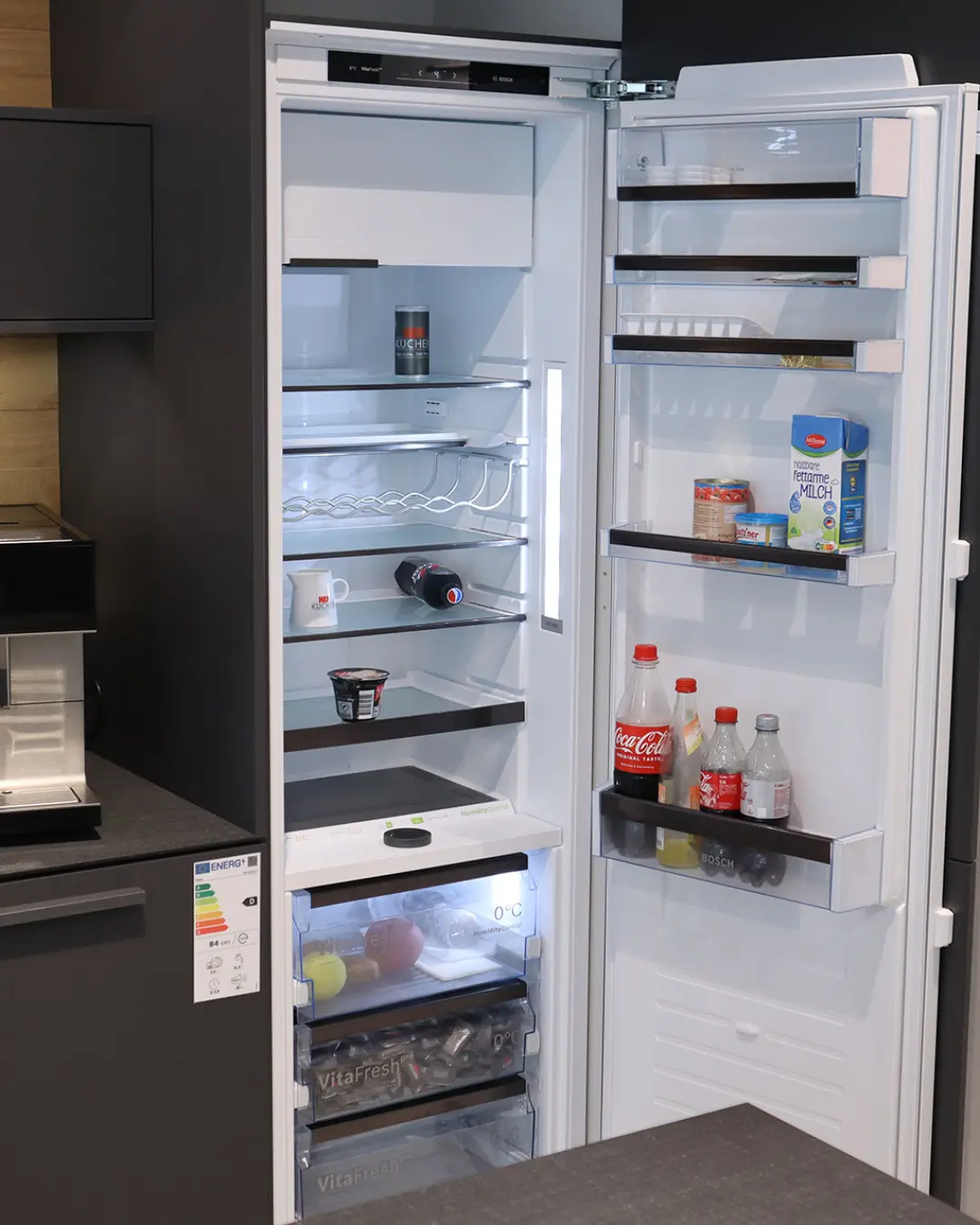 Geöffneter Einbaukühlschrank mit gefüllten Türfächern in der carbongrauen Leicht Küche mit Kochinsel