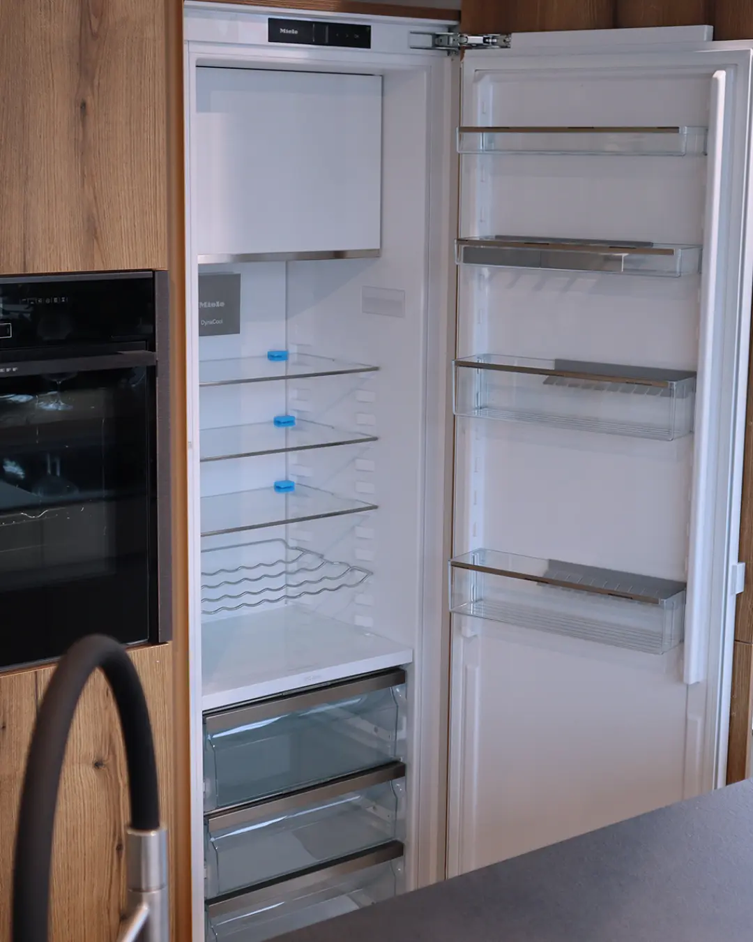 Geöffneter Einbaukühlschrank mit leeren Glasablagen in der grifflosen weißen Leicht Küche KBK19, daneben Backofen in Holzoptik-Front