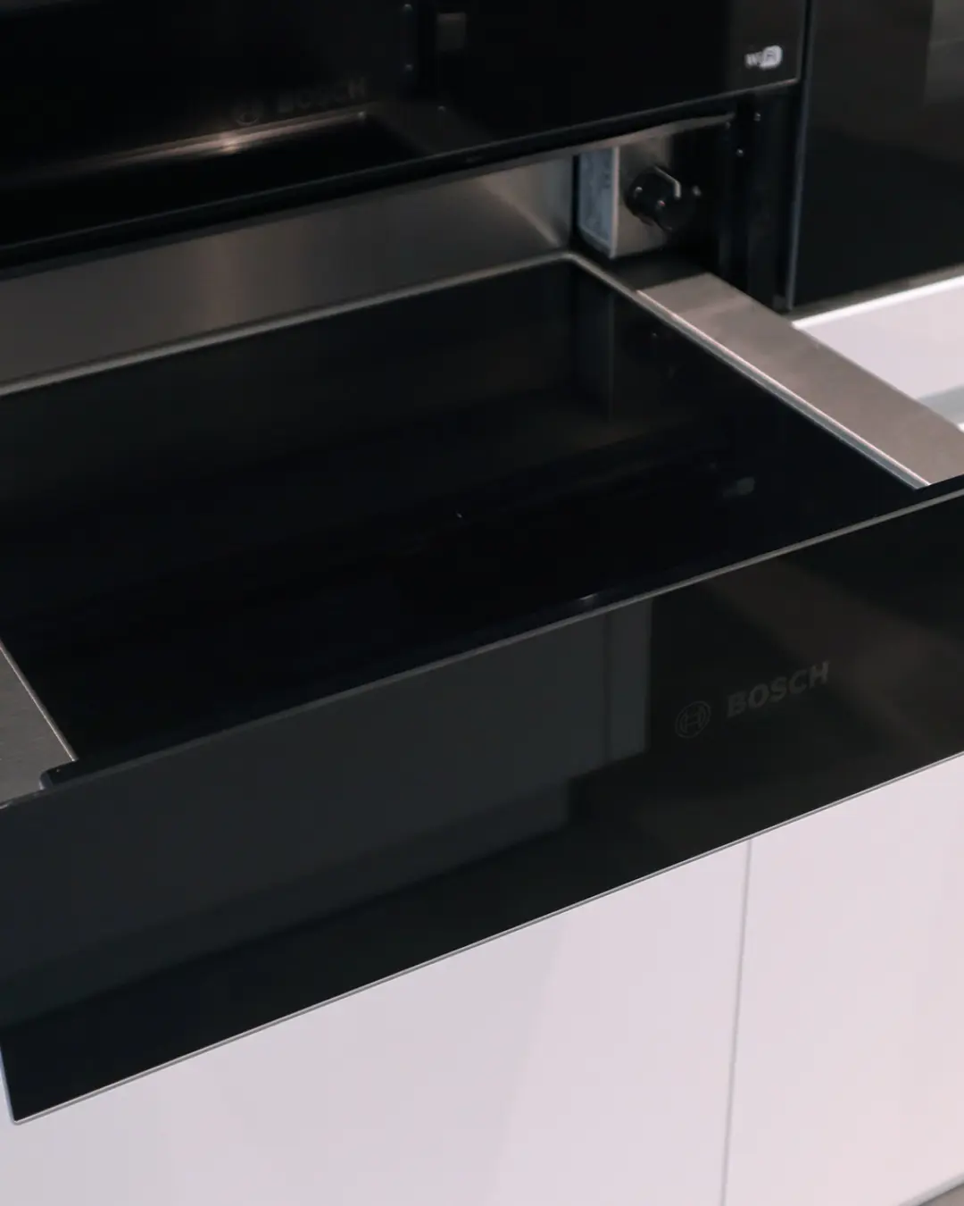 Geöffnete schwarze Wärmeschublade von Bosch in weißer Häcker Küche, darüber ein Einbaugerät
