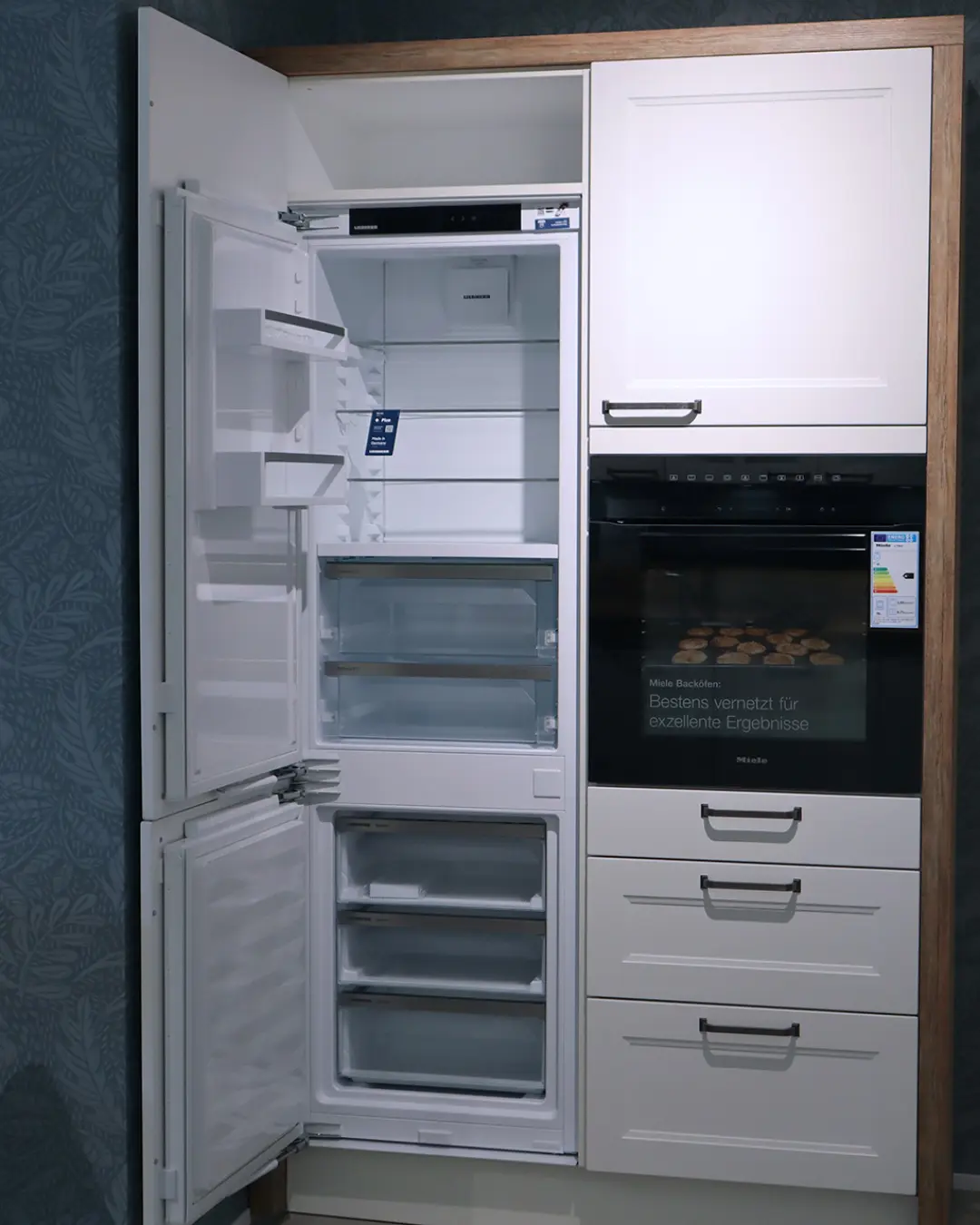 Hochschrankzeile der weißen Landhausküche von Häcker mit geöffnetem Einbaukühlschrank, Backofen und Schubladen