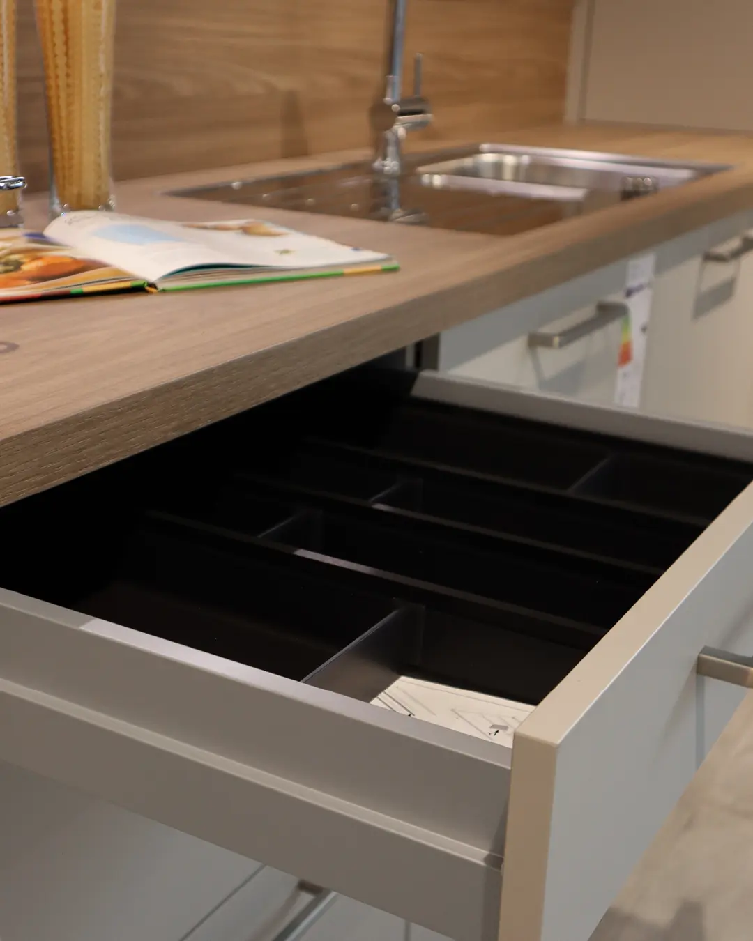 Geöffnete Besteckschublade mit schwarzer Einteilung der Leicht Küche in Kaschmir Beige, darüber Spüle und Arbeitsplatte in Holzoptik
