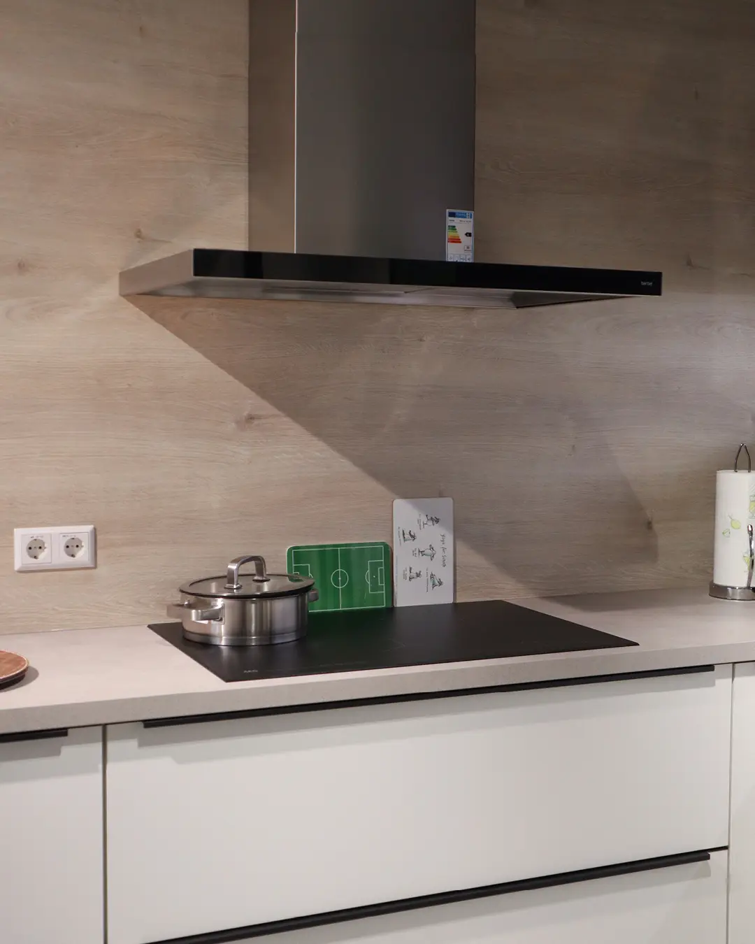 Weiße Nobilia Küche in L-Form: Kochfeld mit Topf, schwarze Wandhaube und Rückwand in Holzoptik