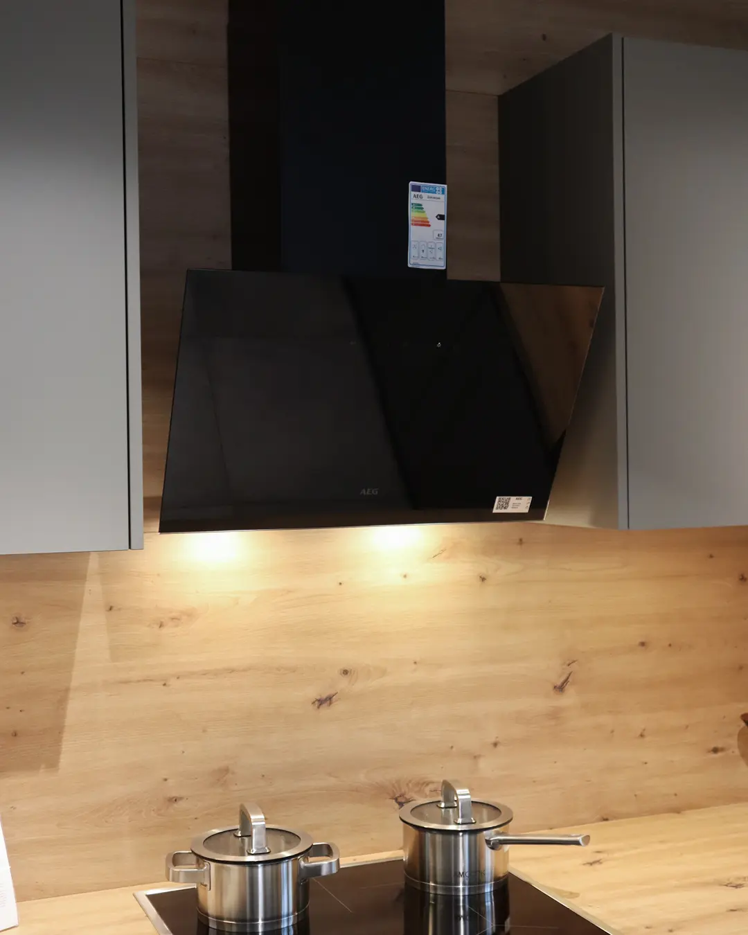 Schwarze Schräghaube über Kochfeld mit Edelstahltöpfen in grauer Nobilia Küche in L-Form, Rückwand in Holzoptik