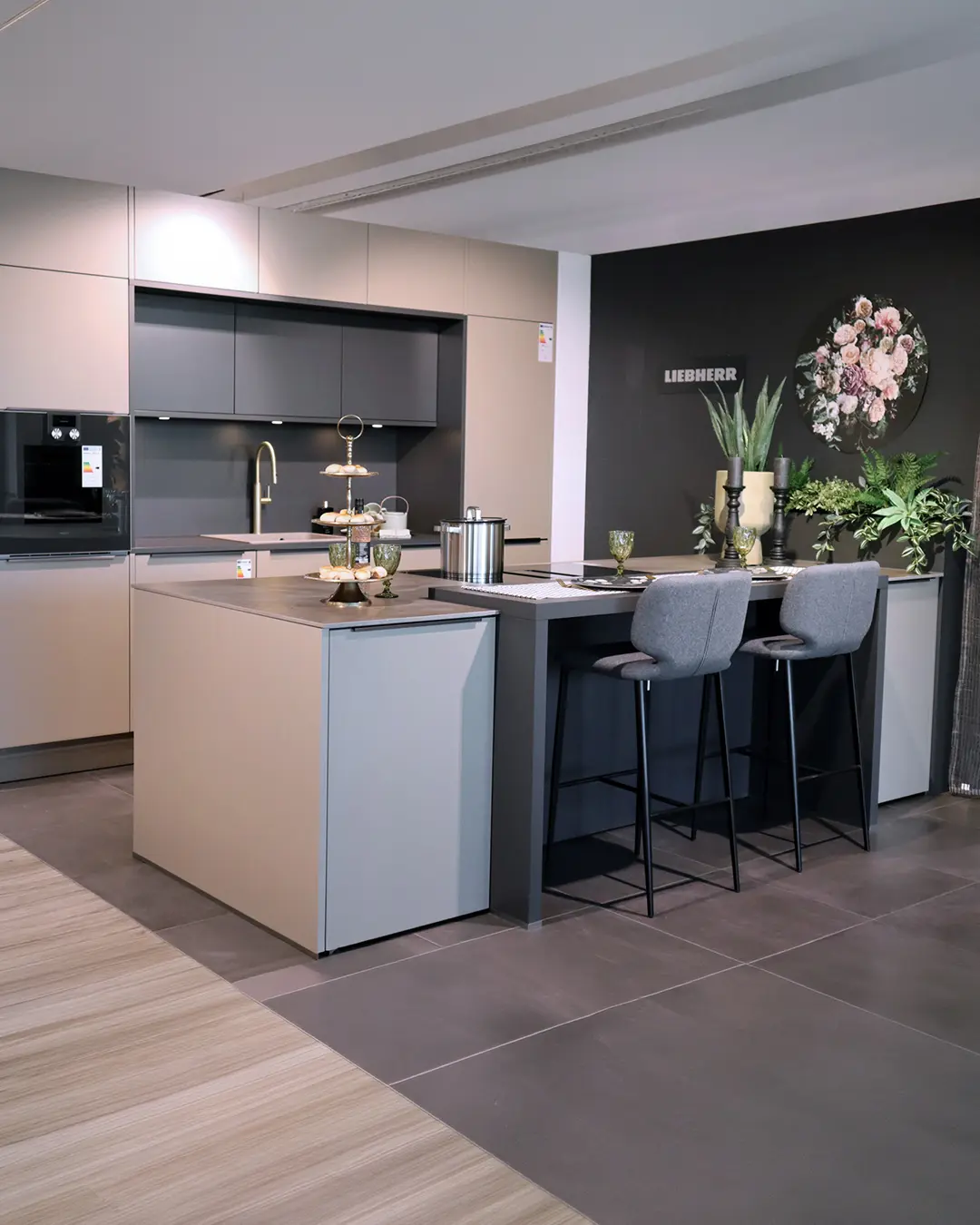 Kaschmir-beige Häcker Küche KCR14 mit Kochinsel, Theke mit zwei grauen Barstühlen und Schriftzug Liebherr an der dunklen Wand