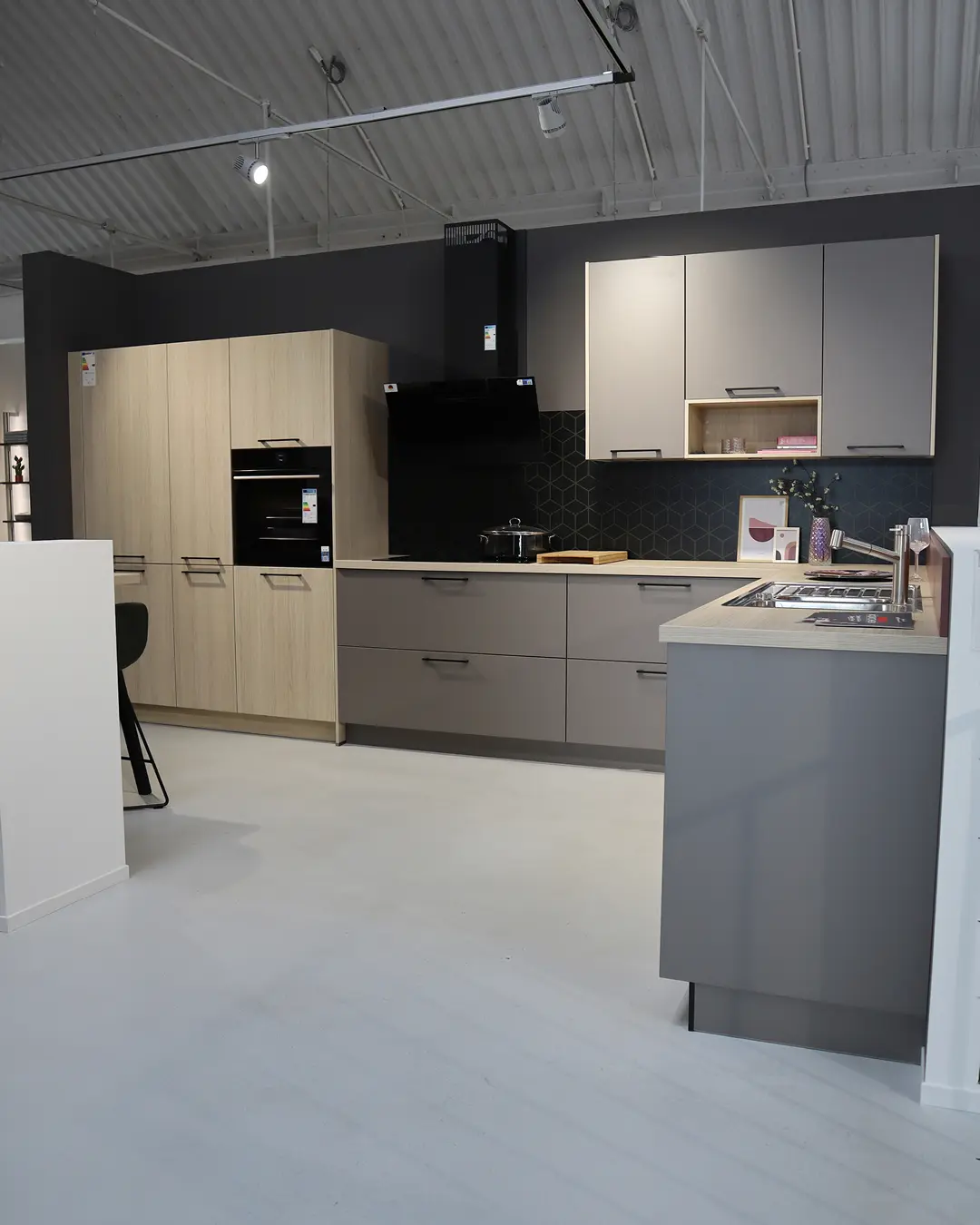 Greige Häcker Küche in L-Form mit Hochschränken in Holzoptik, Kochinsel und dunkler Rückwand im Ausstellungsraum