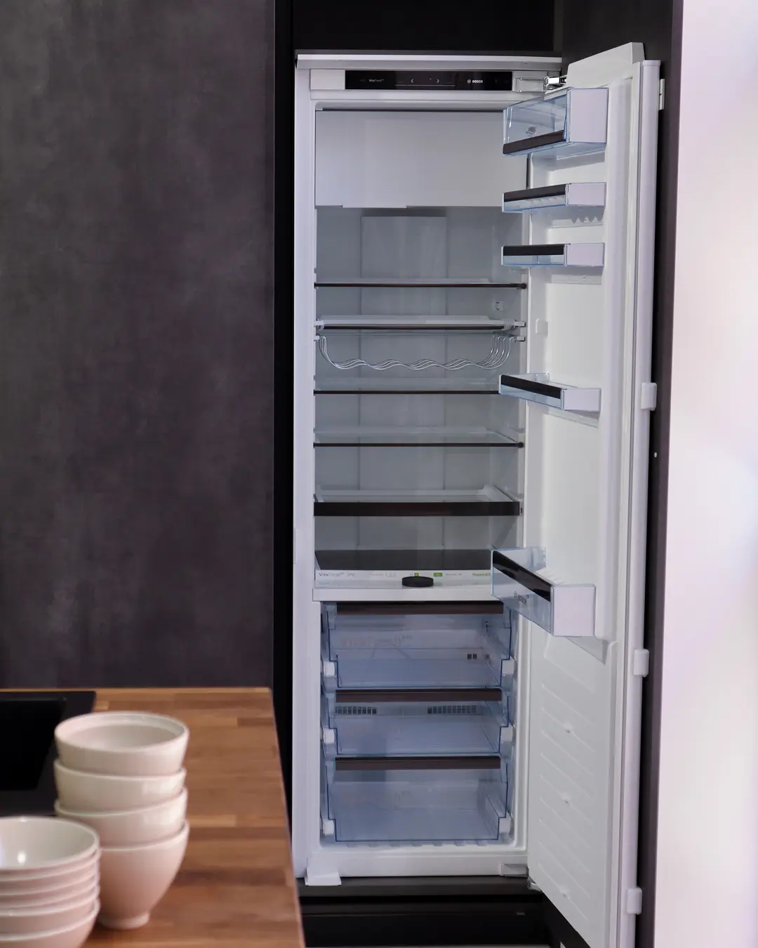 Geöffneter Einbaukühlschrank mit leeren Glasablagen in der Häcker Küche, davor Schalenstapel auf der Arbeitsplatte