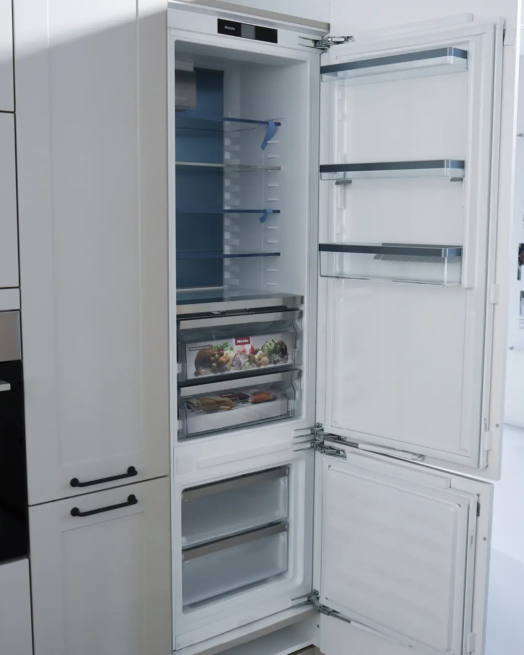 Geöffneter Einbaukühlschrank von Miele mit leeren Glasablagen in der grauen Landhausküche von Häcker