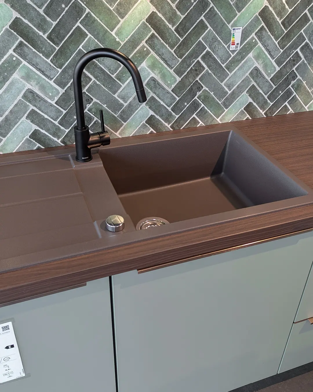 Detail der olivgrünen Nobilia-Küche in U-Form: brauner Spülbecken mit schwarzer Armatur, Arbeitsplatte in Holzoptik und grüner Fischgrätfliesenspiegel
