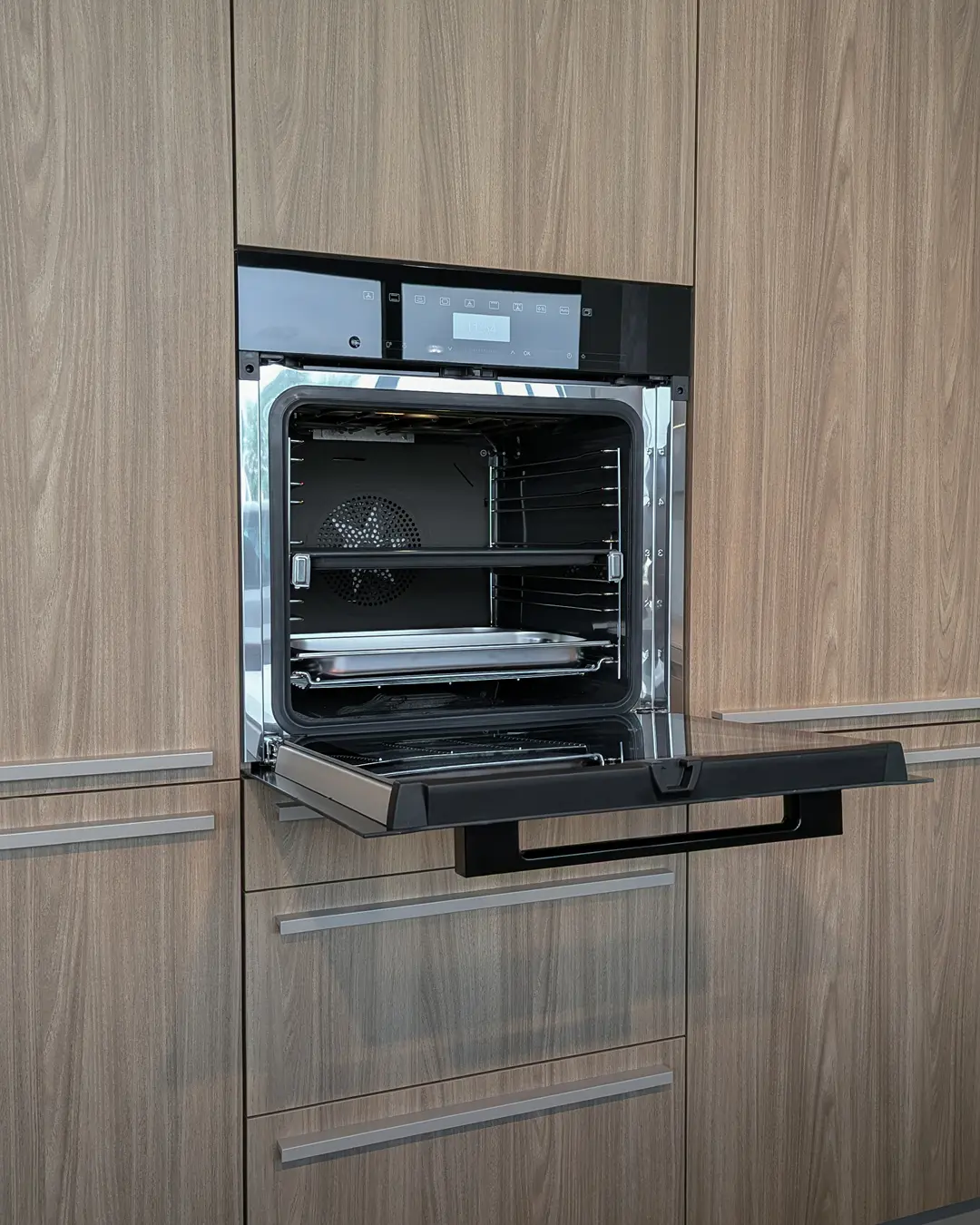 Geöffneter Einbaubackofen in Schwarz mit Backblech, eingebaut in eine Hochschrankfront in Holzoptik