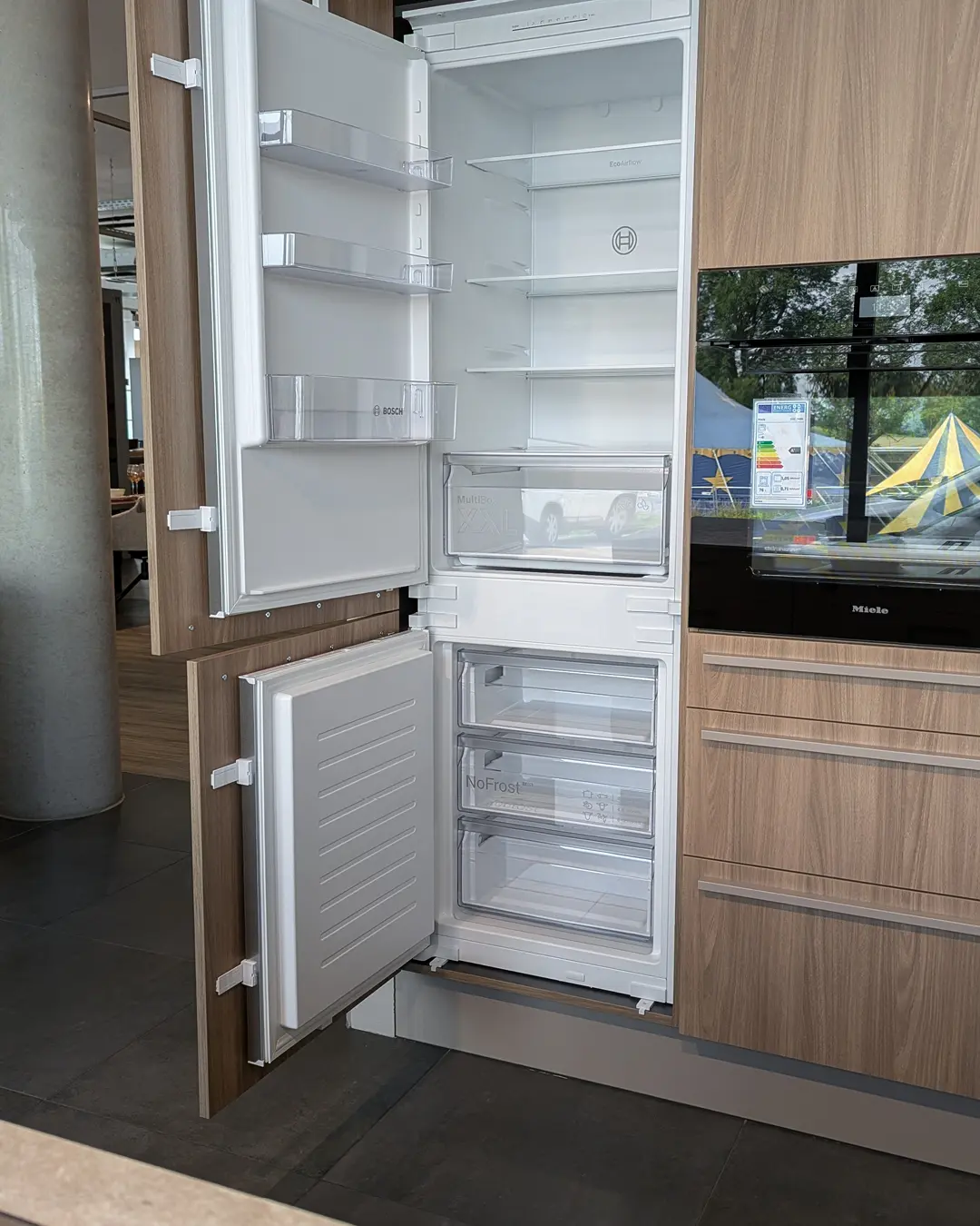 Geöffneter Bosch-Einbaukühlschrank mit NoFrost-Gefrierteil in Holzoptik-Hochschrank neben Miele-Backofen.