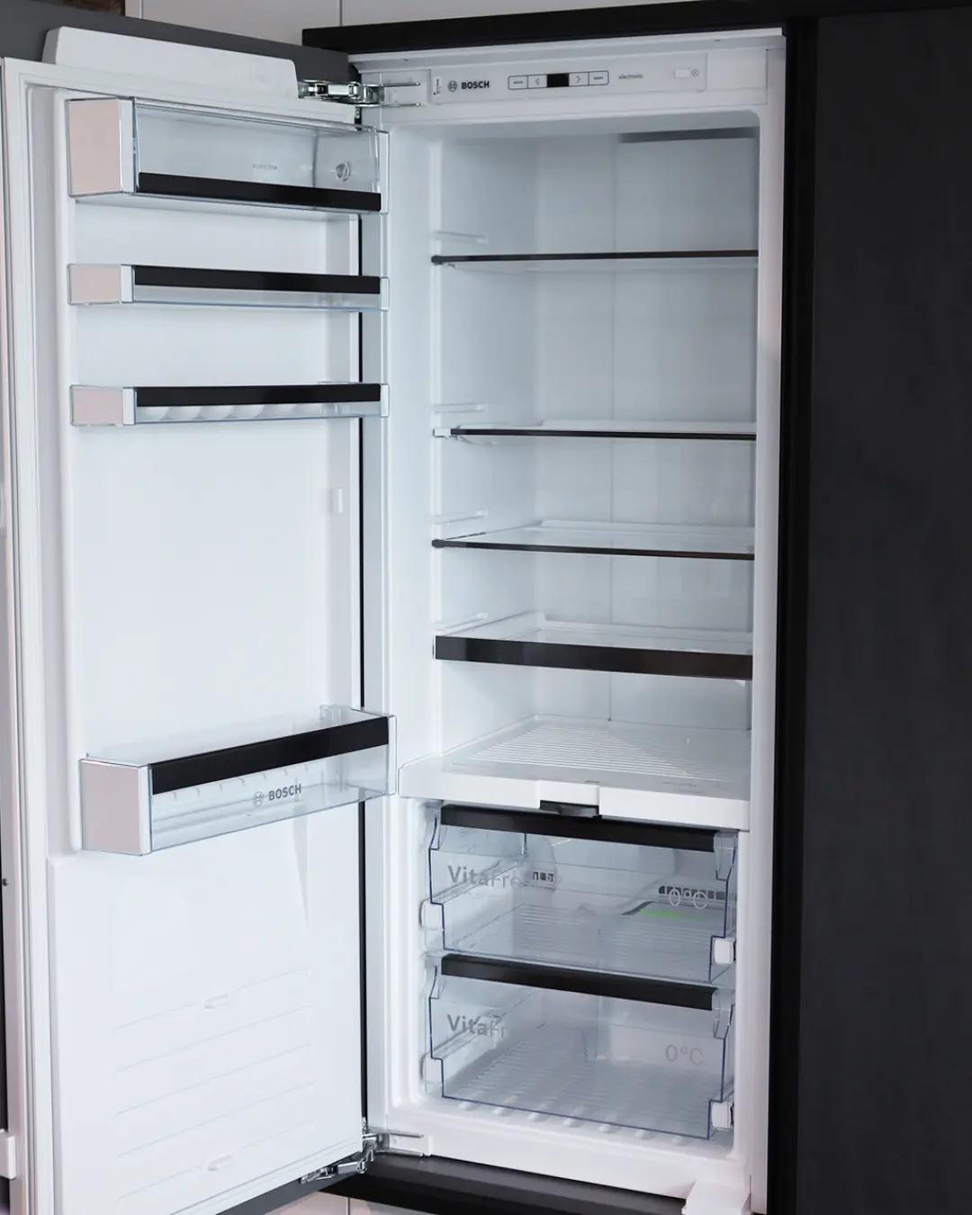 Geöffneter Einbaukühlschrank von Bosch mit leeren Glasablagen und VitaFresh-Schubladen in der weißen Häcker Küche